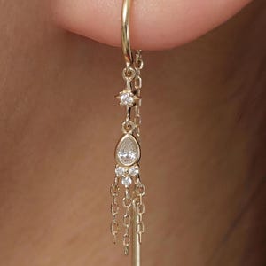 May include: A gold threader earring featuring a delicate chain design with a central pear-shaped clear stone, a small star-shaped accent, and three tiny round clear stones, finished with a smooth gold bar at the end of the dangling chain.