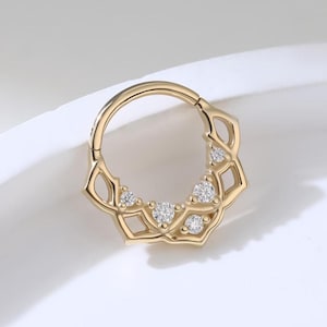 May include: A gold septum ring with a floral design. The ring features small, diamond-like stones set within the petals. The design is symmetrical and the ring is shown against a white background.