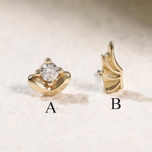 May include: Two gold-toned stud earrings labeled A and B. Earring A features a small, clear, round-cut gemstone set in a curved gold base. Earring B is shaped like a small, textured wing with defined lines, crafted from a gold-toned metal material.