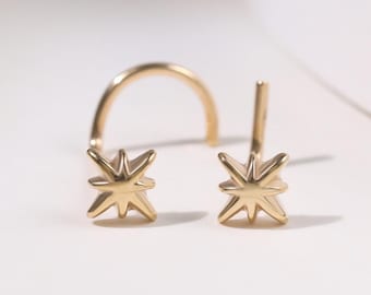 May include: A pair of gold-colored starburst stud earrings and a gold nose ring are displayed on a white surface. The earrings have a star-like design with multiple points. The nose ring is a simple, curved design.