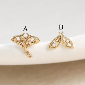 Op de afbeelding: Twee goudkleurige metalen oorknopjes. Oorknopje A heeft de vorm van een mantarog met ingewikkeld opengewerkt patroon. Oorknopje B heeft de vorm van een nachtvlinder met twee kleine heldere steentjes in de vleugels. Gelabeld met zwarte letters A en B.