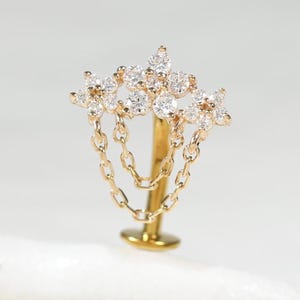 Pode incluir: Um brinco de pino com parte traseira plana e um topo decorativo com cinco pedras claras de corte redondo dispostas em forma de estrela. Duas correntes douradas delicadas pendem do conjunto, conectando-se ao pino central do brinco.