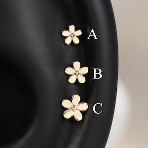 Op de afbeelding: Drie goudkleurige bloemvormige oorknopjes verticaal gerangschikt op een zwart oormodel. Elke oorknop heeft vijf afgeronde bloemblaadjes en een stip in het midden. De oorknopjes zijn gelabeld met de letters A, B en C voor drie verschillende maten.