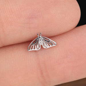 May include: A small, silver-colored butterfly-shaped earring. The earring has a delicate, open-work design, with the wings spread wide. The earring is shown on a finger, against a dark background.