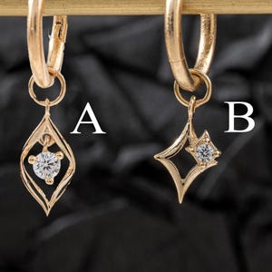 Pode incluir: Dois brincos de argola dourados com pingentes pendurados. Um brinco tem um pingente em forma de gota com um diamante central, rotulado como "A". O outro brinco tem um pingente em forma de estrela com um diamante central, rotulado como "B". Os brincos são exibidos em uma barra dourada sobre um fundo escuro.
