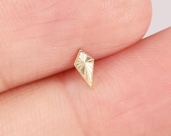 Könnte beinhalten: Ein kleiner, goldfarbener Ohrstecker in geometrischer Drachenform. Der Ohrstecker hat eine strukturierte Oberfläche und wird zwischen zwei Fingern vor einem hellen Hintergrund gehalten. Der Ohrring ist wahrscheinlich für ein einzelnes Piercing.