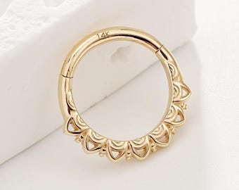 May include: A gold-colored, hinged septum ring with a decorative, scalloped edge. The ring is circular and features a small clasp. The metal is shiny and reflects light. The ring is set against a white background.