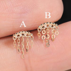 May include: Two small, gold-colored earrings with a fringe design. Each earring features a curved top with small beads and dangling chains. The earrings are displayed on a neutral background.