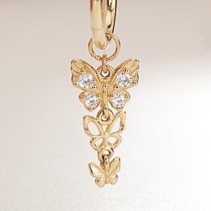 May include: A gold hoop earring featuring a vertical drop charm composed of three connected butterfly shapes. The top butterfly is larger and inlaid with two clear, sparkling stones, while the two smaller butterflies below are plain gold.