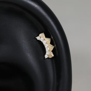 May include: A gold-toned curved ear cartilage piercing stud featuring a fan-like design with five small, clear, round-cut stones set along the inner edge. The jewelry is positioned on a black ear model, showcasing its arc shape and metallic finish.