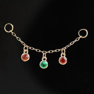 Puede incluir: Un delicado collar de oro con tres colgantes de piedras preciosas de colores: dos rojos y uno verde. El collar se exhibe sobre un cuadrado negro, con un tema festivo de vacaciones en el fondo.