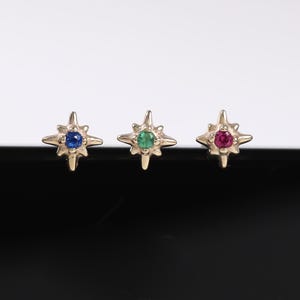 Peut inclure: Trois boucles d'oreilles à clous en forme d'étoile dorée, chacune avec une pierre précieuse de couleur différente au centre : bleue, verte et rouge. Les boucles d'oreilles sont présentées sur un fond noir et blanc.