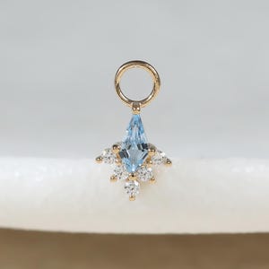May include: A gold charm featuring a central marquise-cut light blue gemstone accented by a cluster of small, clear round-cut stones at the base. The charm includes a circular jump ring at the top for attachment to a chain or bracelet.