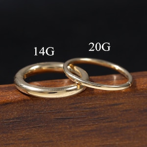 Peut inclure: Deux anneaux circulaires de couleur dorée avec des épaisseurs différentes. Le plus grand anneau est marqué 14G et le plus petit, plus fin, est marqué 20G. Les deux anneaux ont une finition lisse et polie et sont posés sur une surface en bois.