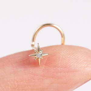 May include: A gold nose ring with a starburst design and a small, clear gemstone. The ring has a curved post. The jewellery is shown against a plain white background. The starburst is a delicate, five-pointed shape.