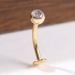 May include: A gold-coloured belly button ring with a small, clear gemstone at the top. The jewellery has a curved bar and a flat disc at the bottom. The background is a mix of white and brown.