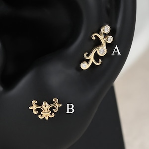 Peut inclure: Deux boucles d'oreilles dorées présentées sur un modèle d'oreille noir. La boucle A est une pièce incurvée en forme de vigne avec trois petites pierres claires. La boucle B est un motif symétrique inspiré de la fleur de lys.