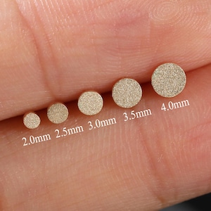 May include: A collection of six small, round, gold-colored stud earrings in graduated sizes, ranging from 2.0mm to 4.0mm in diameter. The earrings are displayed on a light-toned background, showcasing their subtle sparkle and varying dimensions.