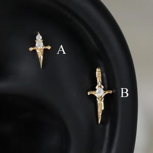 May include: Two gold-toned sword-shaped earrings labeled A and B. Earring A features a small, pointed blade with a single clear stone at the hilt. Earring B is a longer, detailed sword design with a clear stone set in the center of the crossguard.