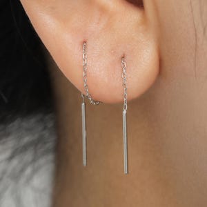 Può includere: Un paio di orecchini a barra argentati. Ogni orecchino presenta una delicata catena collegata a una barra dritta e sottile. Gli orecchini sono progettati per essere indossati attraverso l'orecchio, creando un look minimalista e moderno.