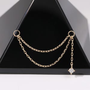 May include: A gold-colored earring with a chain design. The earring features two small hoops connected by delicate chains, with a small, square-cut gemstone dangling at the end. The earring is displayed against a black, triangular object.