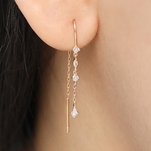 May include: A gold-colored threader earring with three clear, sparkling gemstones. The earring is worn in the ear and hangs down, with a small bar at the end. The gemstones are different shapes and sizes.