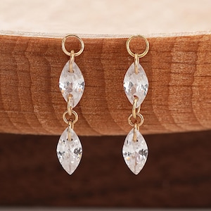 Puede incluir: Un par de pendientes colgantes de tono dorado con dos piedras transparentes de corte marquesa suspendidas verticalmente de un pequeño aro circular. Cada pendiente consta de dos cristales facetados unidos por finos conectores de metal dorado.
