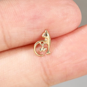 May include: A small gold-toned stud earring shaped like a stylized cat. The cat figure is depicted in a seated position with its tail curved upward, holding a tiny dangling star-shaped charm within the loop of its tail. The metal has a polished finish.