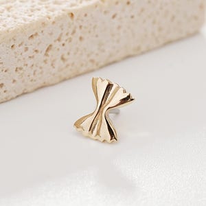 Pode incluir: Um brinco de pressão dourado em forma de laço. O brinco é pequeno e detalhado, com uma superfície texturizada. O brinco está sobre um fundo branco com um bloco bege texturizado ao fundo.