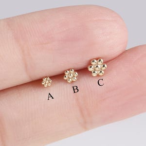 May include: Three small, gold-colored flower-shaped stud earrings. The earrings are labeled A, B, and C, with varying sizes. The earrings are displayed on a light-colored surface.
