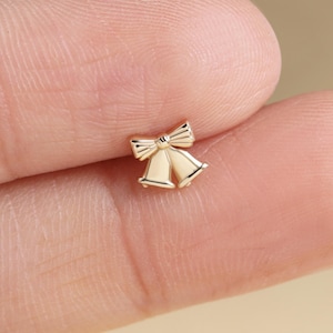 May include: A small, gold-colored Christmas bell charm with a bow on top. The charm is held between two fingers, with a blurred background featuring Christmas decorations.