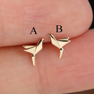 Puede incluir: Dos pequeños pendientes de botón dorados con forma de pájaros de origami. El pendiente de la izquierda está etiquetado con la letra A y el de la derecha con la letra B. Cada pájaro presenta líneas geométricas de estilo papel plegado.