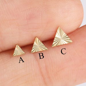 May include: Three small, gold-colored triangle-shaped earrings. Each earring has a different textured design. The earrings are labeled A, B, and C. The earrings are displayed on a light-colored background.