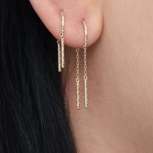 May include: A close-up of two gold earrings. One earring features a small hoop with a dangling bar, while the other has a chain with a bar. The earrings are worn on a person's ear, with the focus on the jewelry.