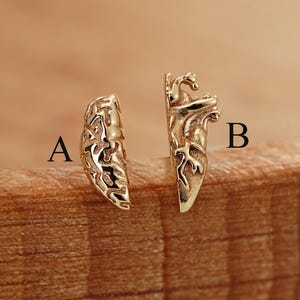 May include: Two gold-colored earrings are displayed on a wooden surface. One earring is a curved, open design with intricate patterns. The other earring has a more detailed, three-dimensional shape with a similar gold finish. The earrings are labeled A and B.