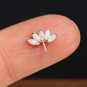 May include: A small, gold-colored earring with a floral design. The earring features five white, oval-shaped gemstones arranged in a petal-like pattern. The post is silver-colored. The earring is held between two fingers against a blurred background.