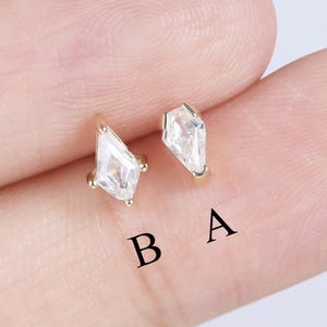 May include: Two small, gold-colored earrings with a geometric, crystal-like design. The earrings are held between fingers, with the letters "B" and "A" visible below them.