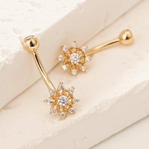 Könnte beinhalten: Ein Paar goldfarbene Ohrringe mit floralem Design. Jeder Ohrring hat einen zentralen klaren Stein, der von kleineren klaren Steinen umgeben ist und einen Sterneneffekt erzeugt. Die Ohrringe liegen auf einer weißen Oberfläche.