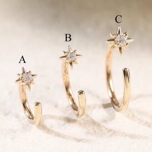 May include: Three gold hoop earrings labeled A, B, and C, each featuring a star-shaped charm set with a clear gemstone. The hoops are open-ended, and the star charms are positioned at the top of each hoop, displaying a metallic gold finish.