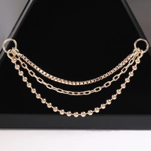 May include: A gold necklace with three fine chains is suspended in front of a black pyramid. The chains feature a box chain, a curb chain, and a beaded chain. The jewellery is displayed against a white backdrop.
