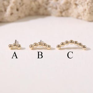 Puede incluir: Se muestran tres pendientes de botón dorados. El primero tiene tres pequeñas cuentas, el segundo tiene cinco y el tercero tiene siete cuentas en forma de arco. Los pendientes están etiquetados A, B y C.