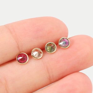 Puede incluir: Cuatro pequeños pendientes de oro con piedras preciosas redondas y de colores. Los pendientes están dispuestos en fila en un dedo, con la piedra roja a la izquierda y la piedra verde a la derecha.