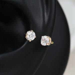 May include: Two round, brilliant-cut clear stones set in gold-toned metal prongs. Each stone is secured in a stud-style earring setting, featuring a polished metallic post and back, designed to be worn as a pair of earrings on the earlobe.