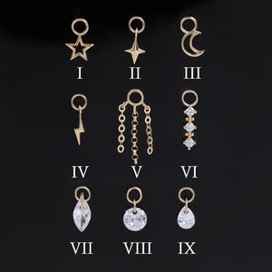 May include: Nine gold-coloured charms with diverse designs, presented on a black surface. The charms feature a star, teardrop, moon, lightning bolt, chain, and various crystal shapes. Each charm is labelled with a Roman numeral.