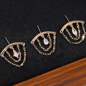 Peut inclure: Trois boucles d'oreilles dorées avec une gemme blanche en forme de goutte d'eau et des détails de chaîne délicats. Les boucles d'oreilles ont un dessus incurvé et sont présentées sur une surface noire dans un cadre en bois. Elles sont pour oreilles percées.