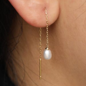 Può includere: Primo piano di un orecchino a catena dorata con una piccola perla bianca. L'orecchino pende dall'orecchio. L'orecchino ha una lunga e sottile barra d'oro da un lato e una catena con una perla dall'altro.