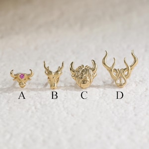 May include: Four gold-toned metal stud earrings labeled A, B, C, and D. Earring A features a small bull head with a pink gemstone. Earring B is a deer head. Earring C is a shaggy highland cow head. Earring D is a stylized deer head with antlers.