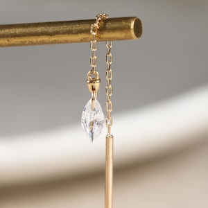 May include: A gold chain threader earring featuring a marquise-cut clear gemstone and a slender gold bar drop. The delicate chain loops over a horizontal gold rod, showcasing a minimalist design with a polished metallic finish and a faceted stone.