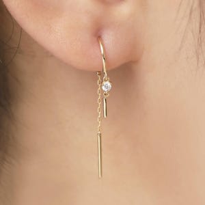 Könnte beinhalten: Ein goldfarbener Ohrring mit einem kleinen, klaren Edelstein und einem herabhängenden Stab. Der Ohrring ist an einem kleinen Bügel befestigt, der durch das Ohr geht. Eine zarte Kette und ein zweiter Stab hängen unter dem Edelstein.