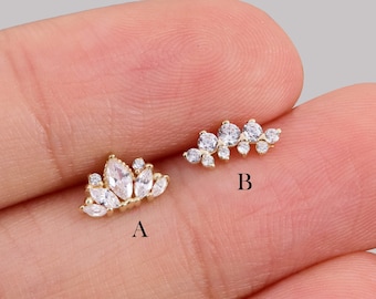 May include: Two gold-toned, small earrings with clear, sparkling stones. One earring is a crown shape, and the other is a cluster of stones. The earrings are displayed on a light-colored background.